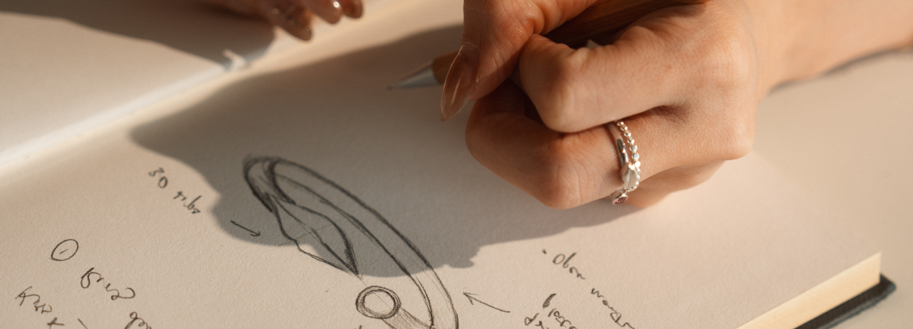 Close-up of a Inari designer's hand drawing a minimalist ring design on paper, adorned with a delicate gemstone for the ring